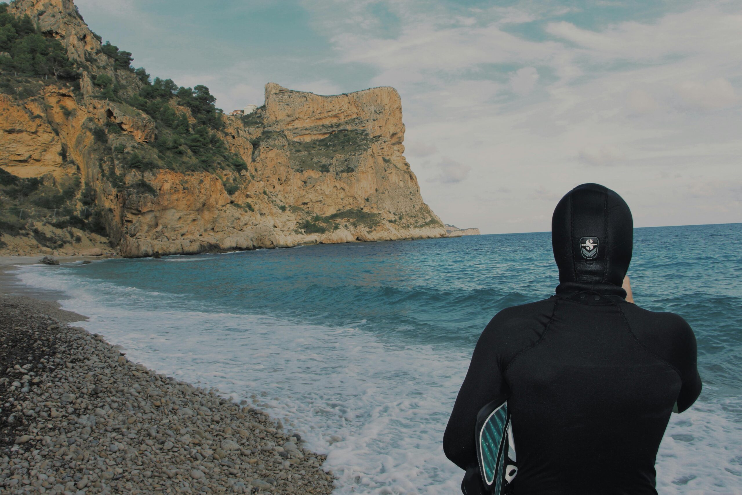 A diver in wetsuit on rocky beach with stunning cliff and ocean waves under a clear sky.